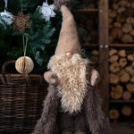 Plush gnome with a long beard and tall hat in front of a decorated Christmas tree and woodpile.