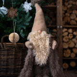 Plush gnome with a long beard and tall hat in front of a decorated Christmas tree and woodpile.