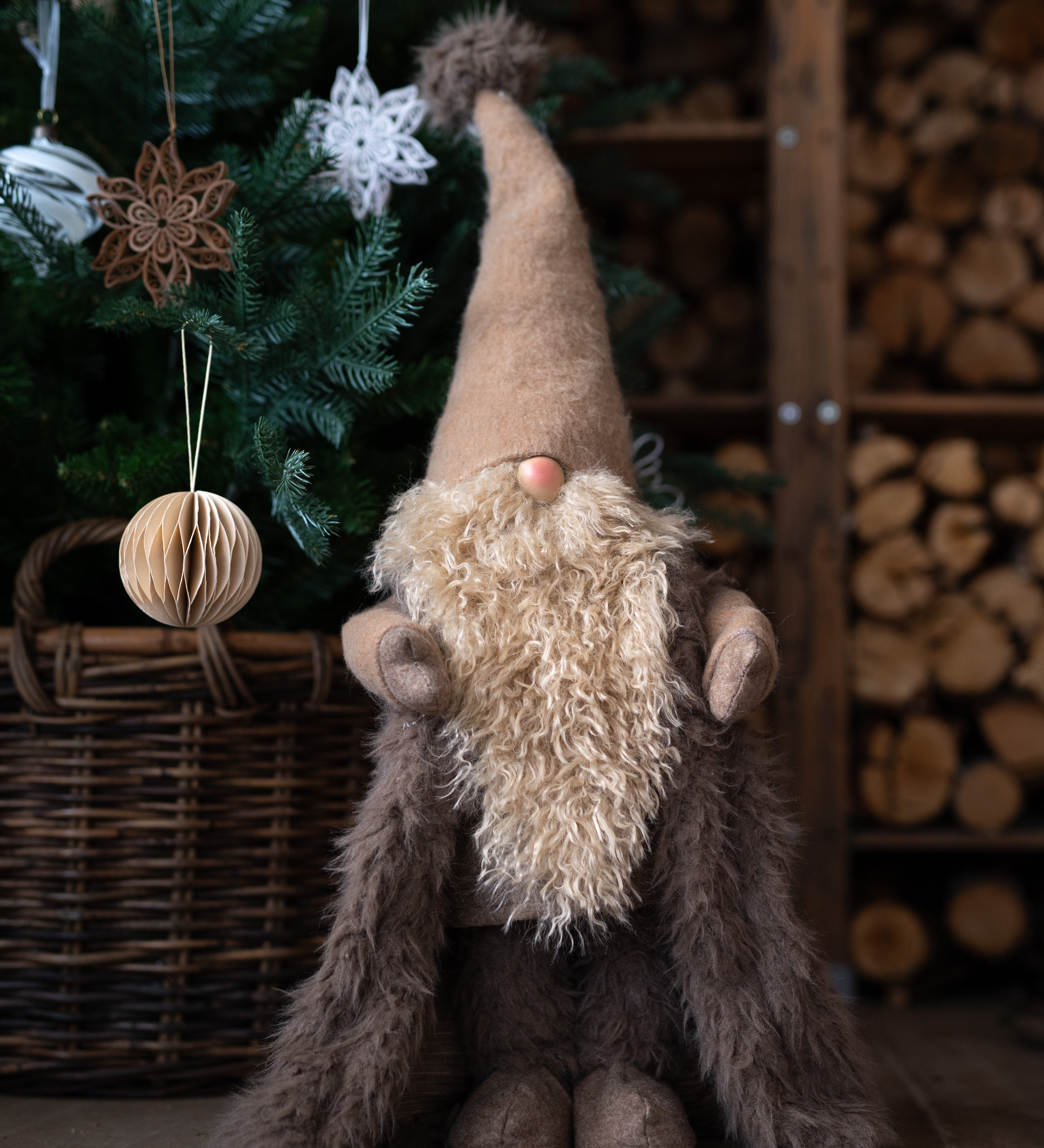 Plush gnome with a long beard and tall hat in front of a decorated Christmas tree and woodpile.