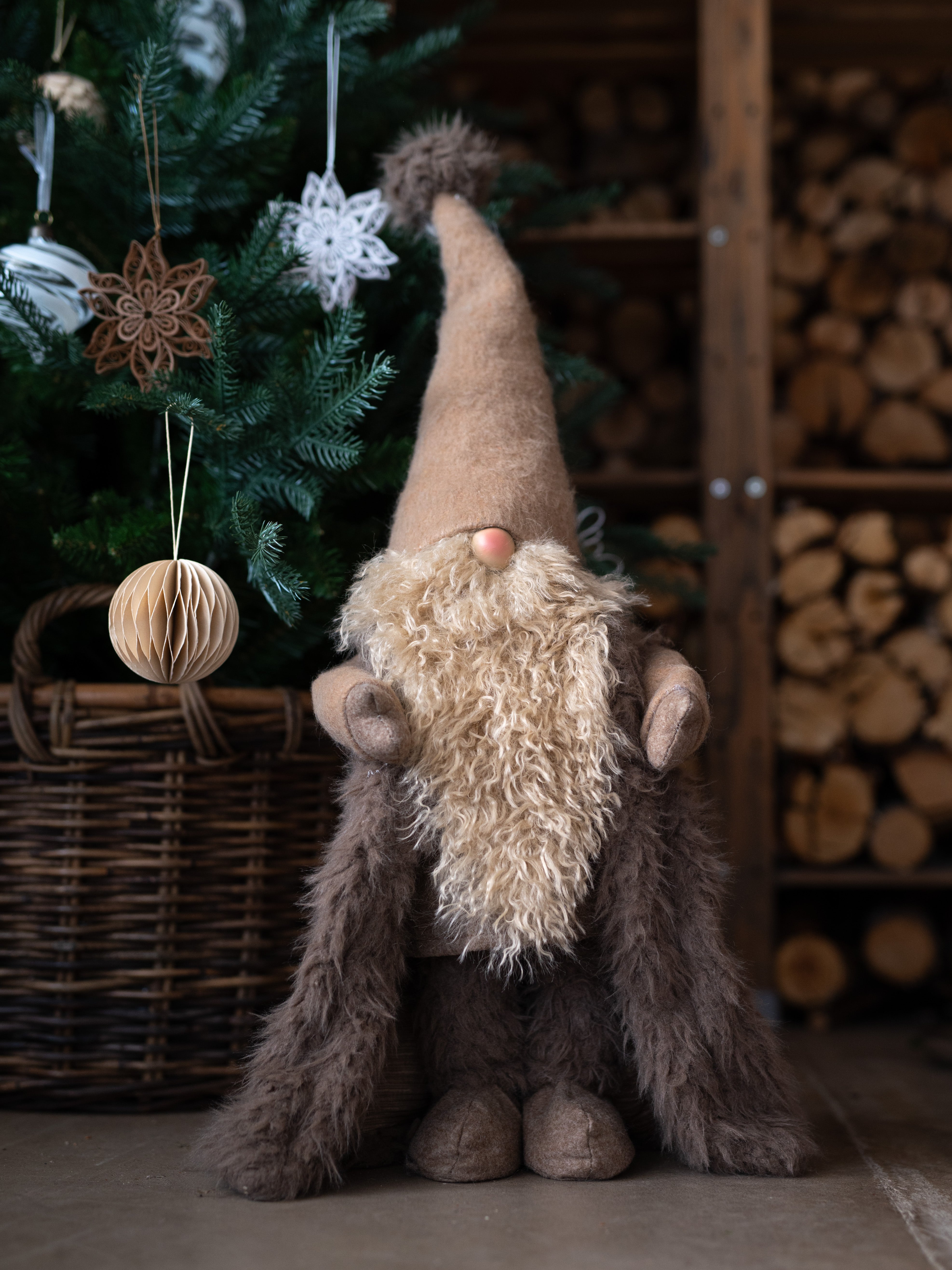 Plush gnome with a long beard and tall hat in front of a decorated Christmas tree and woodpile.